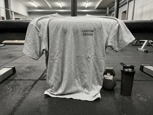 Drop-shoulder custom T-shirt on a gym floor beside a dumbbell, printed with CUSTOM DESIGN